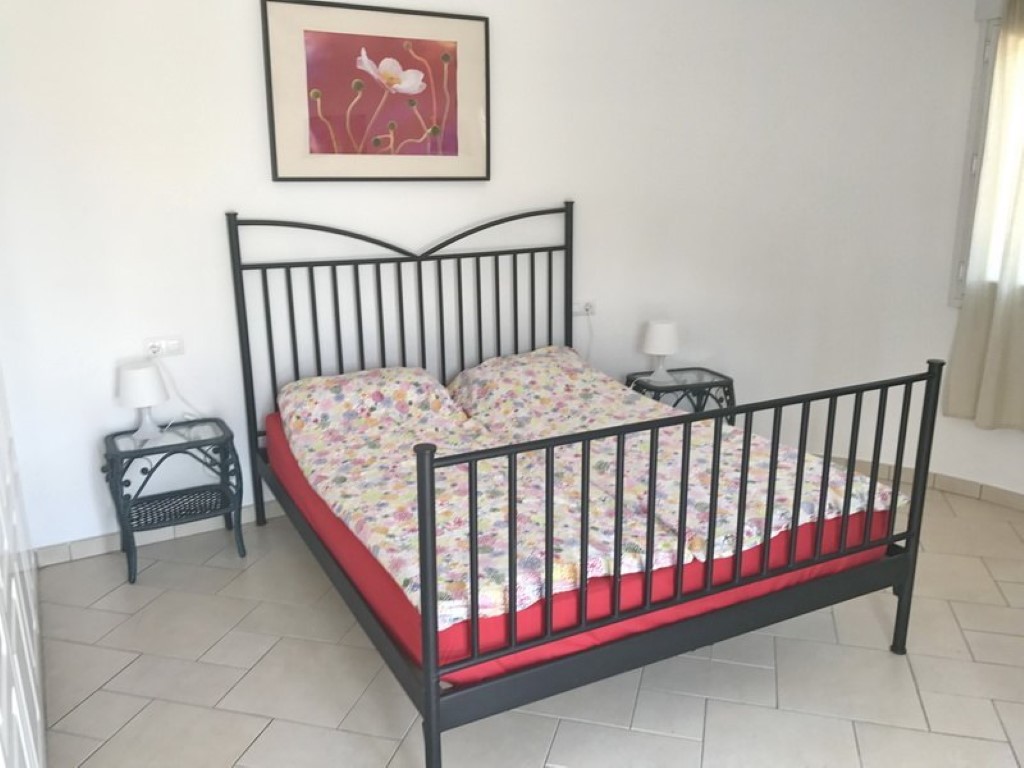 Slaapkamer met een zwart smeedijzeren bedframe, bijpassende nachtkastjes van riet en glas, en een lichte tegelvloer. De ruimte geniet van natuurlijke lichtinval.