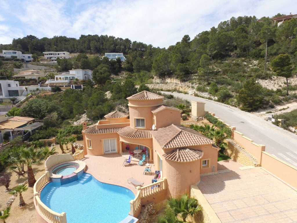 Luchtfoto van de mediterrane villa met gebogen zwembad, ronde jacuzzi en groot zonneterras, gelegen tegen een beboste heuvel in een rustige wijk.