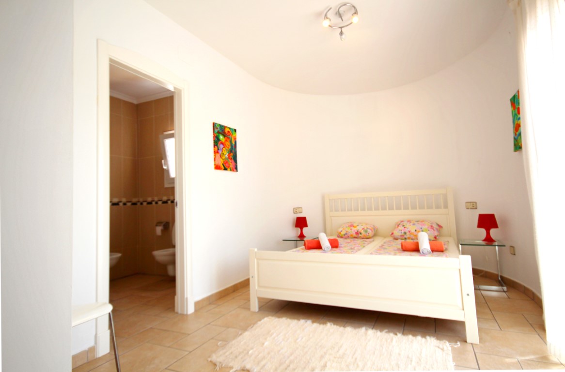 Slaapkamer met een gebogen wand, wit houten bedframe en een aangrenzende badkamer met neutrale tegels.