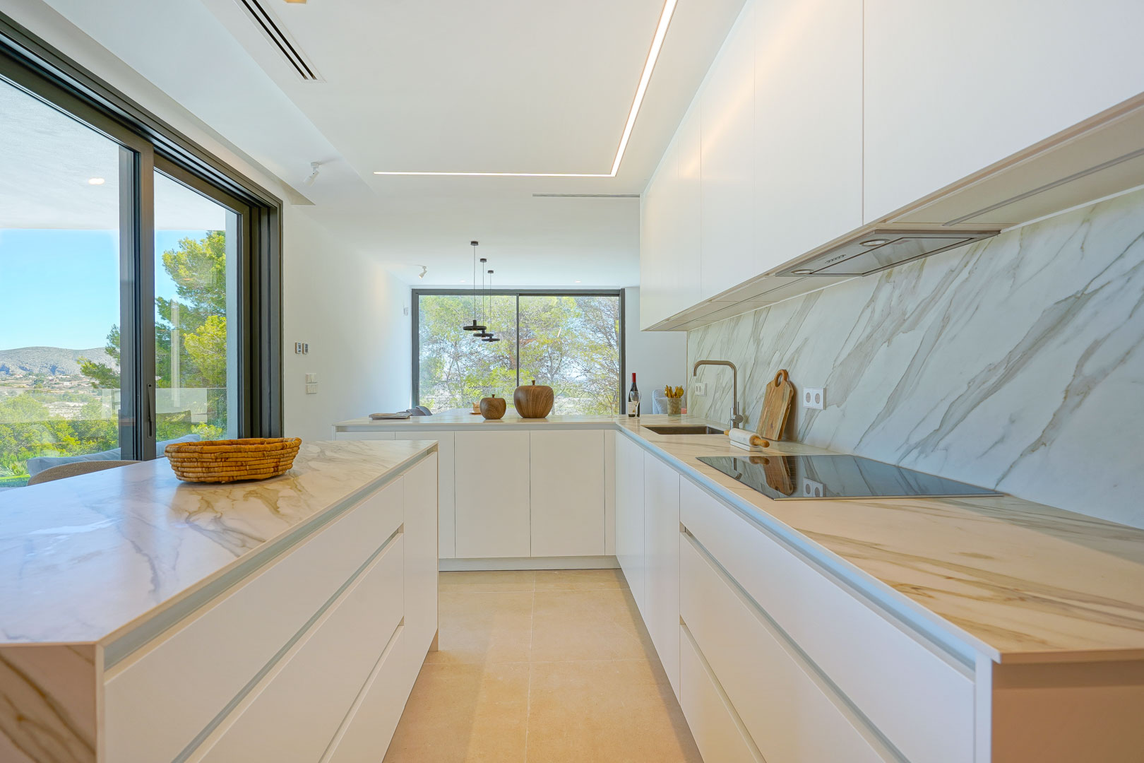 Minimalistische open keuken met marmerlook werkbladen, greeploze witte kasten en lineaire LED-verlichting. Grote raampartijen bieden uitzicht op de bosrijke omgeving.