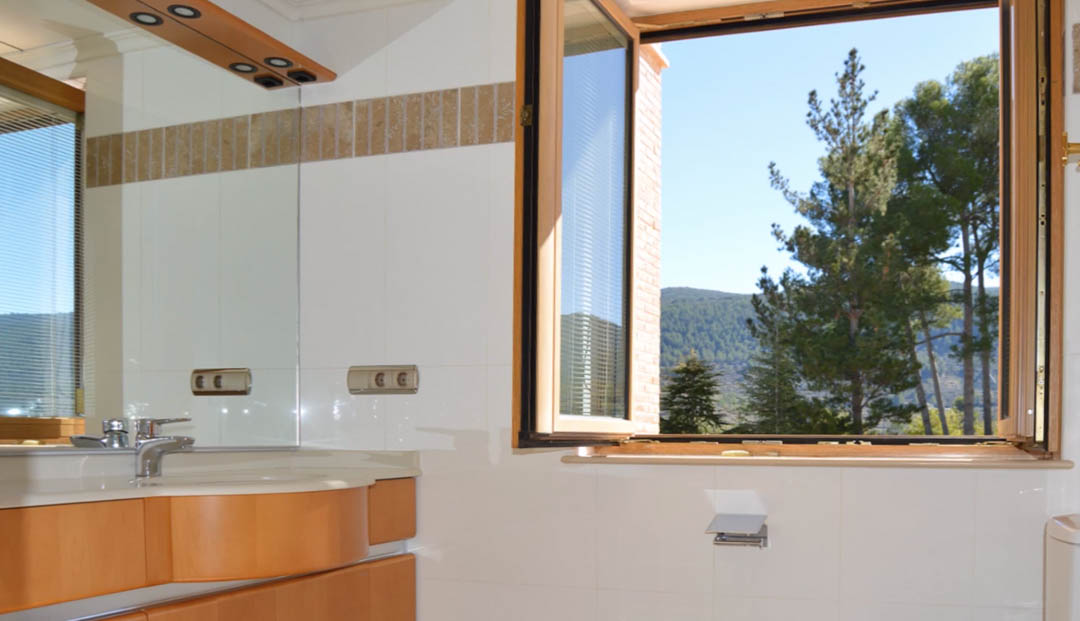 Badkamer met houten wastafelmeubel, grote spiegel en een open raam met uitzicht op de omliggende dennenbossen en het bergachtige landschap.
