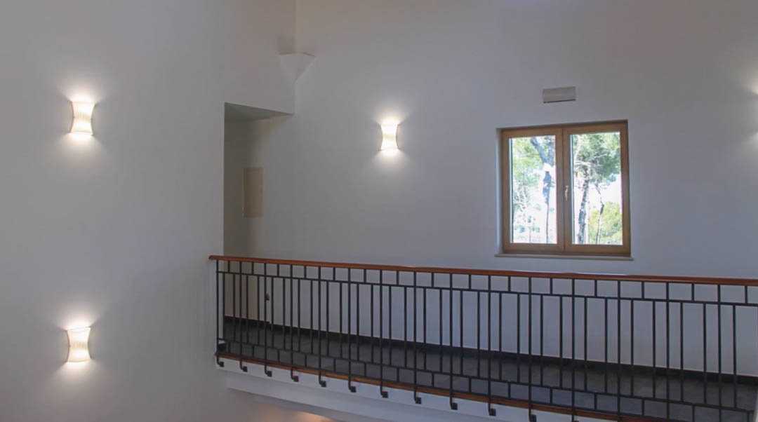 Bovenverdieping met smeedijzeren balustrade en houten handgreep, verlicht door moderne wandlampen en een raam met uitzicht op de natuurlijke omgeving.