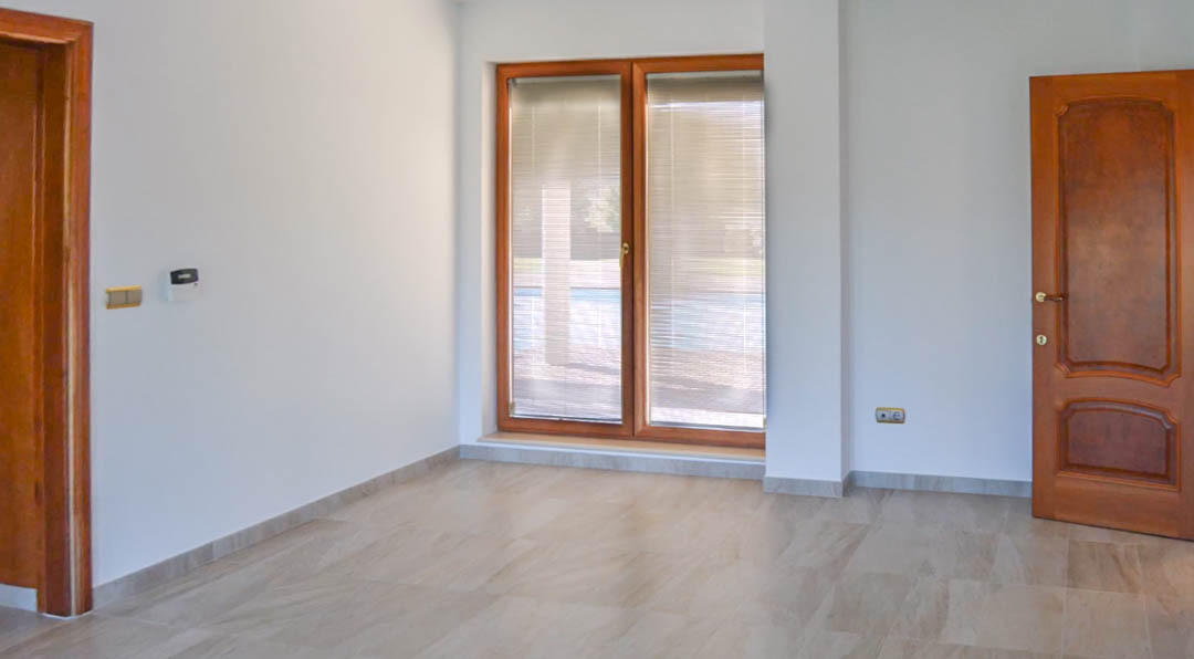 Lichte kamer met natuurstenen vloer, hoogwaardig houten deuren en kozijnen, en zicht op het zwembad via de ramen met geïntegreerde lamellen.