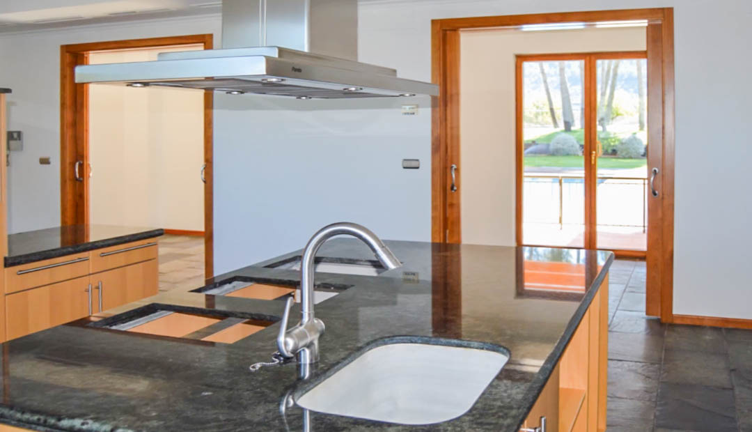 Moderne keuken met een donker granieten kookeiland, rvs afzuigkap en natuurstenen vloer. Grote houten deurkozijnen bieden toegang tot het terras en de tuin.
