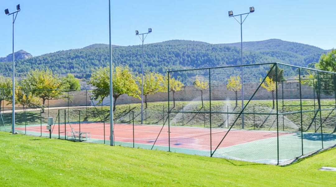 Omheinde tennisbaan met een rood-groen oppervlak, voorzien van hoge lichtmasten en een panoramisch uitzicht op de beboste bergen.