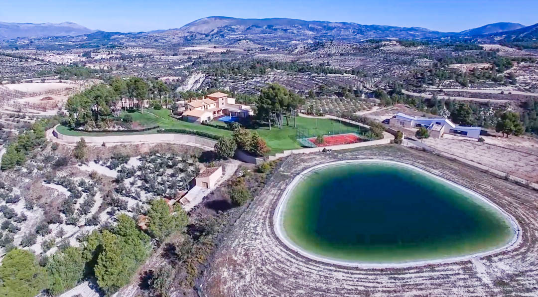 Panoramisch luchtfoto van het landgoed met het grote waterreservoir, de rode tennisbaan en de hoofdvilla, omgeven door uitgestrekte bergen en velden.