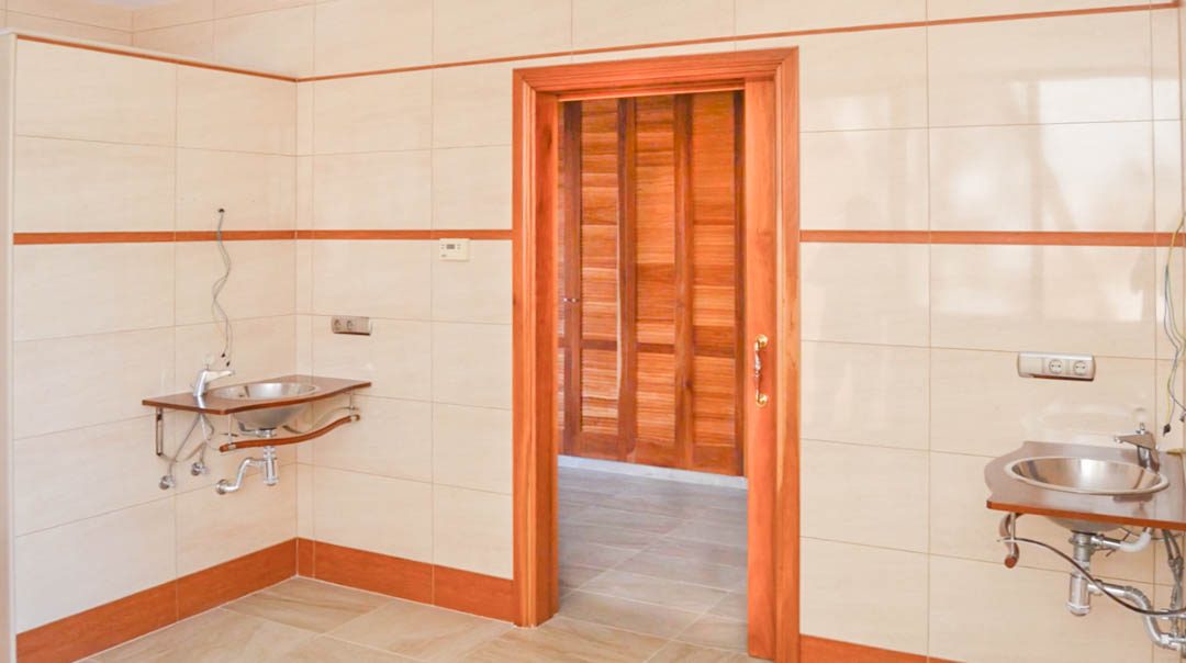 Wasruimte met roestvrijstalen wastafels en betegelde wanden, die toegang biedt tot een kleedkamer met hoogwaardige houten kasten.