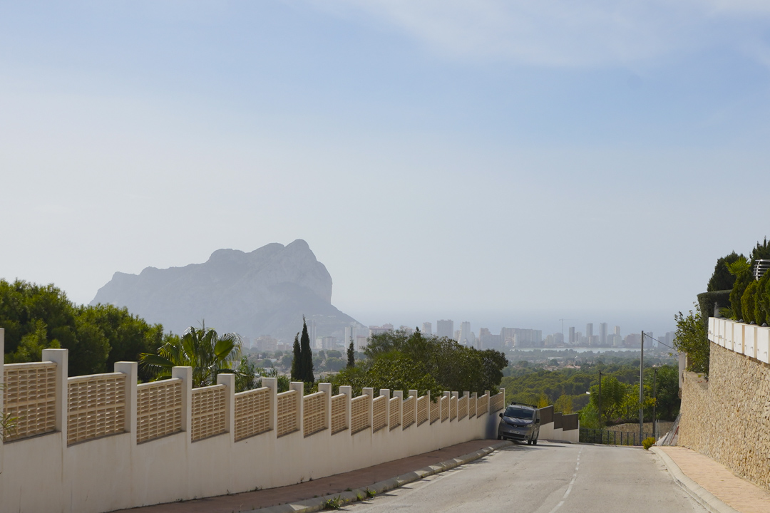 Panoramisch uitzicht vanaf de toegangsweg op de Peñón de Ifach en de skyline van Calpe, geflankeerd door natuurstenen muren en witte hekwerken.