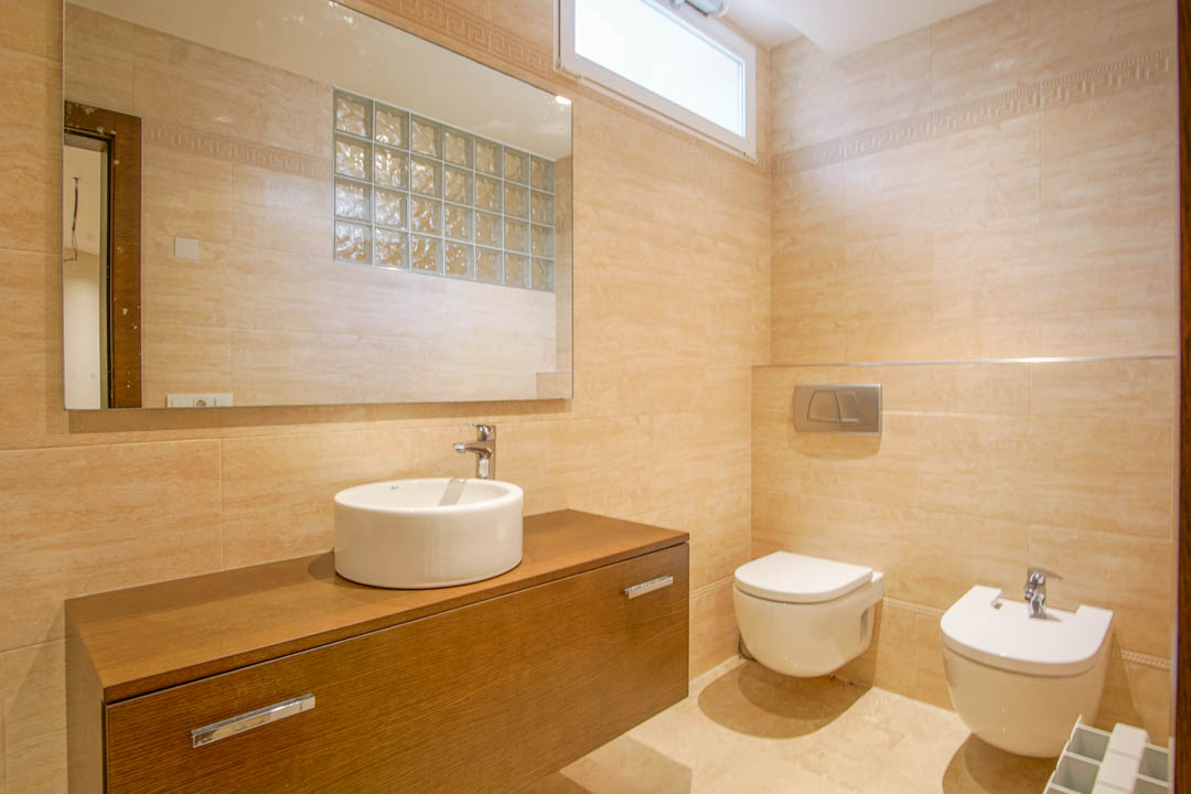 Badkamer met beige betegeling, zwevend houten wastafelmeubel met opzetkom, wandcloset en bidet, en een spiegel die een wand van glasbouwstenen reflecteert.