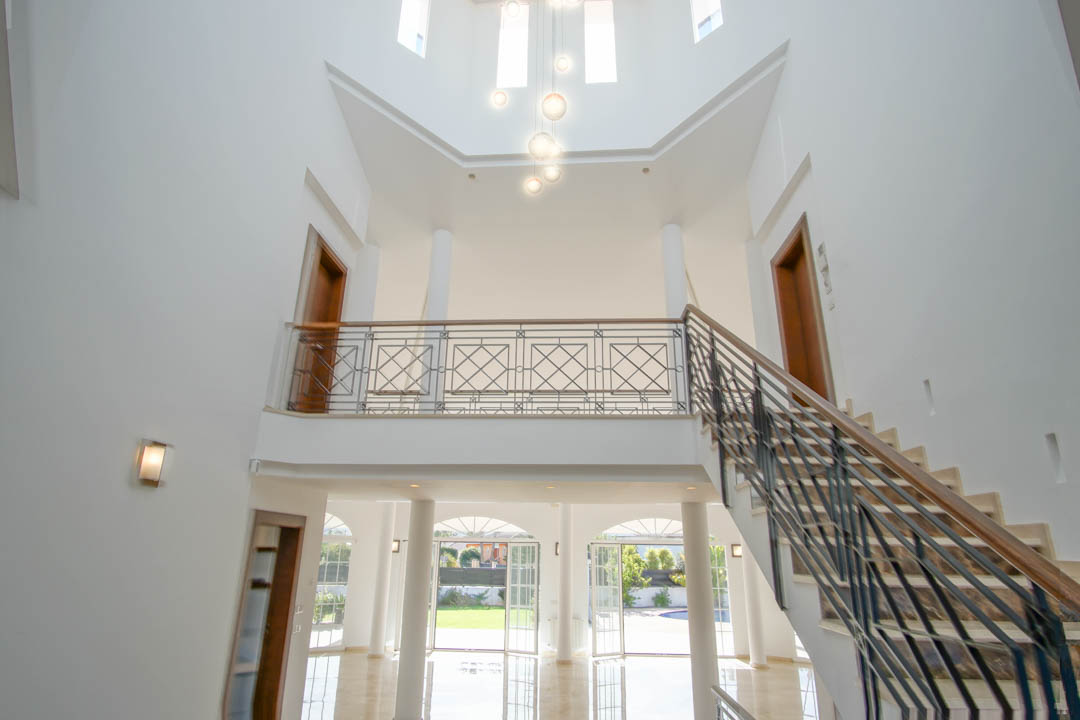 Dubbelhoge hal met marmeren trap, smeedijzeren leuning en een bovenliggende galerij, verlicht door moderne hanglampen en grote openslaande deuren naar de tuin.