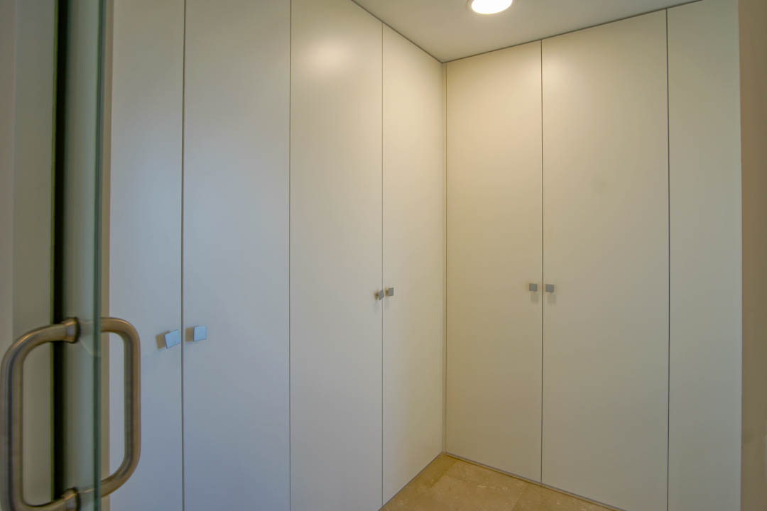 Kamerhoge inbouwkasten in het wit met vierkante metalen handgrepen, natuurstenen vloer en verlichting via inbouwspots.