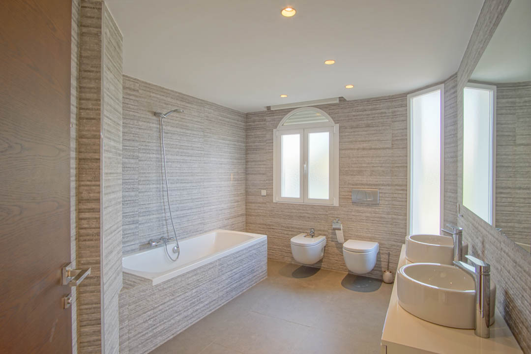 Moderne badkamer met dubbele opzetwastafel, ingebouwd ligbad en getextureerde wandtegels. Voorzien van hangend sanitair en natuurlijk lichtinval via een rondboogvenster.