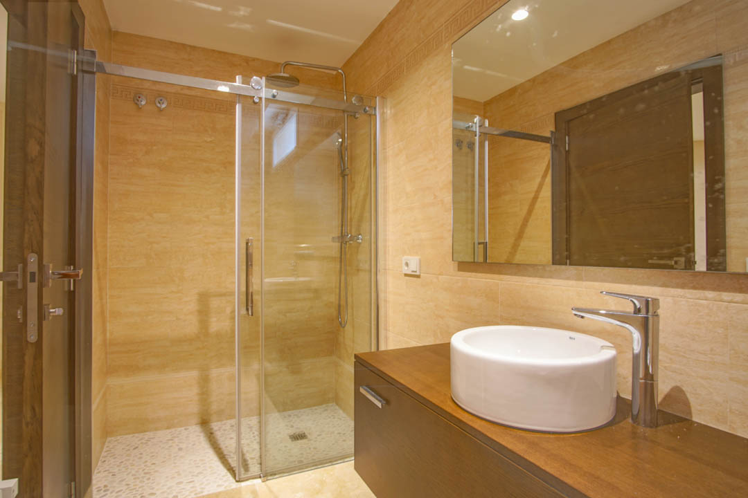 Moderne badkamer met glazen douchecabine, kiezelvloer, ronde opzetwastafel op een donkerhouten meubel en warme beige wandbetegeling.