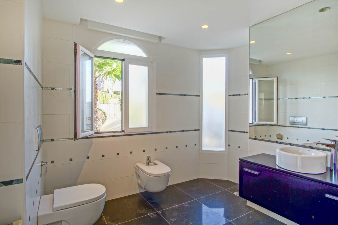 Moderne badkamer met hangend toilet en bidet, donker wastafelmeubel en witte wandtegels. Een boograam zorgt voor natuurlijk licht en uitzicht op de tuin.
