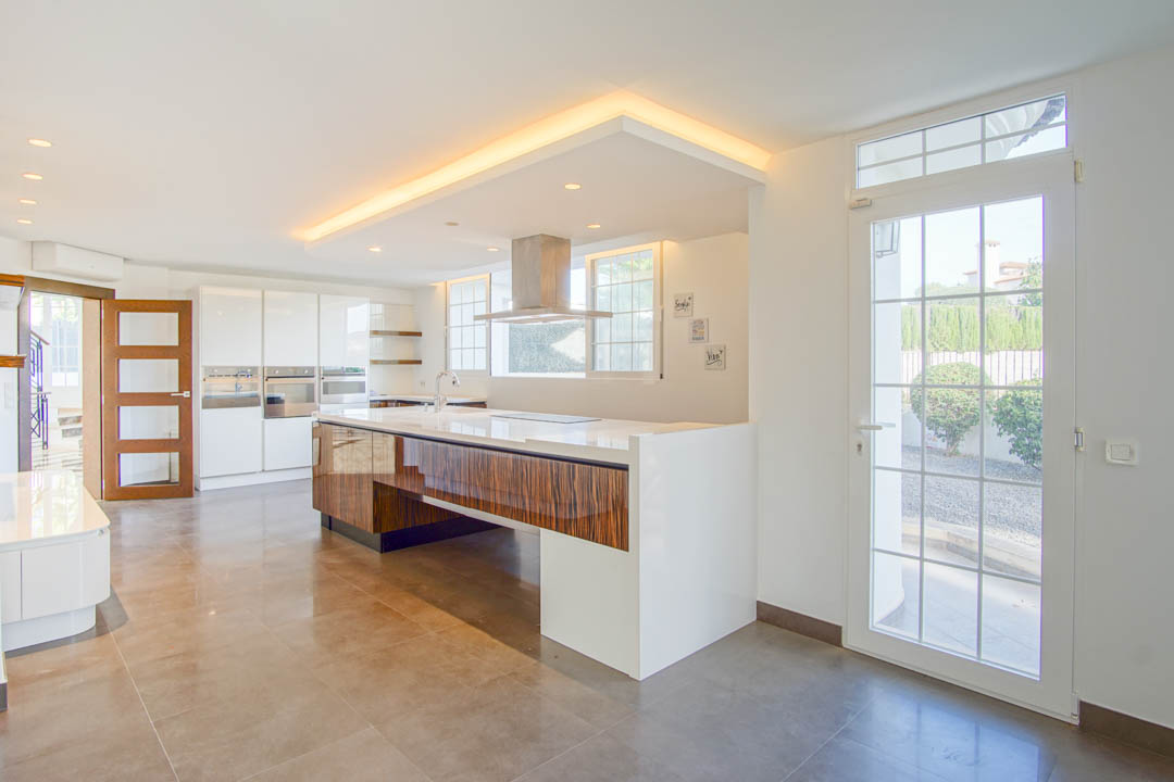 Moderne keuken met een groot kookeiland in houtnerf, wit werkblad en rvs afzuigkap. Voorzien van indirecte plafondverlichting en toegang tot de tuin.