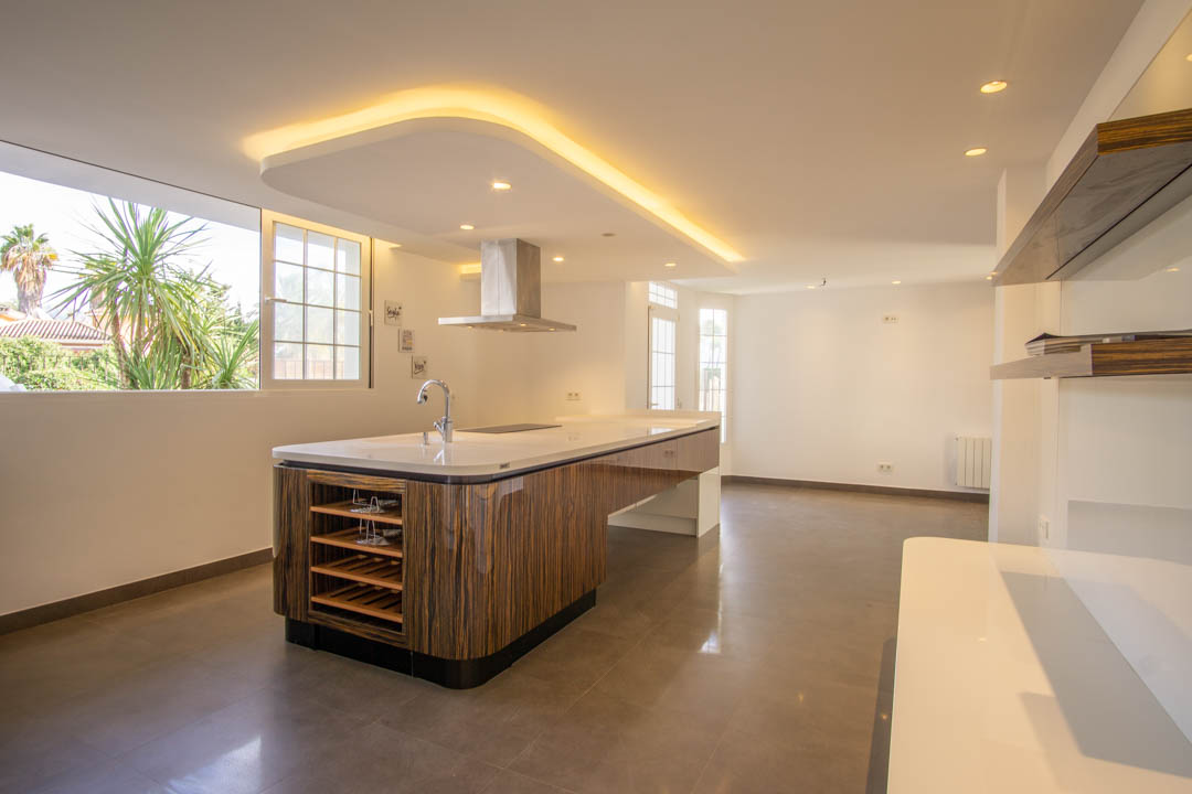 Moderne keuken met een kookeiland in houtnerf-afwerking, wit werkblad en rvs afzuigkap. Voorzien van indirecte led-plafondverlichting en uitzicht op de volgroeide tuin.