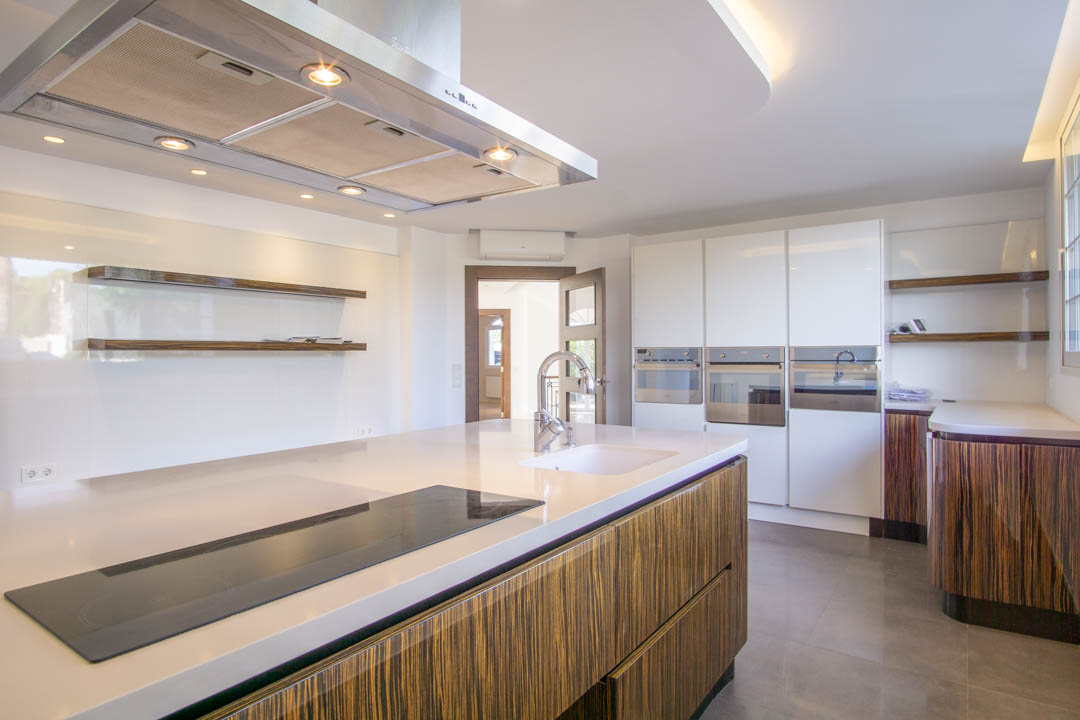 Moderne keuken met kookeiland, wit werkblad en kasten met houtnerf. Voorzien van inbouwapparatuur, een roestvrijstalen afzuigkap en minimalistische zwevende planken.