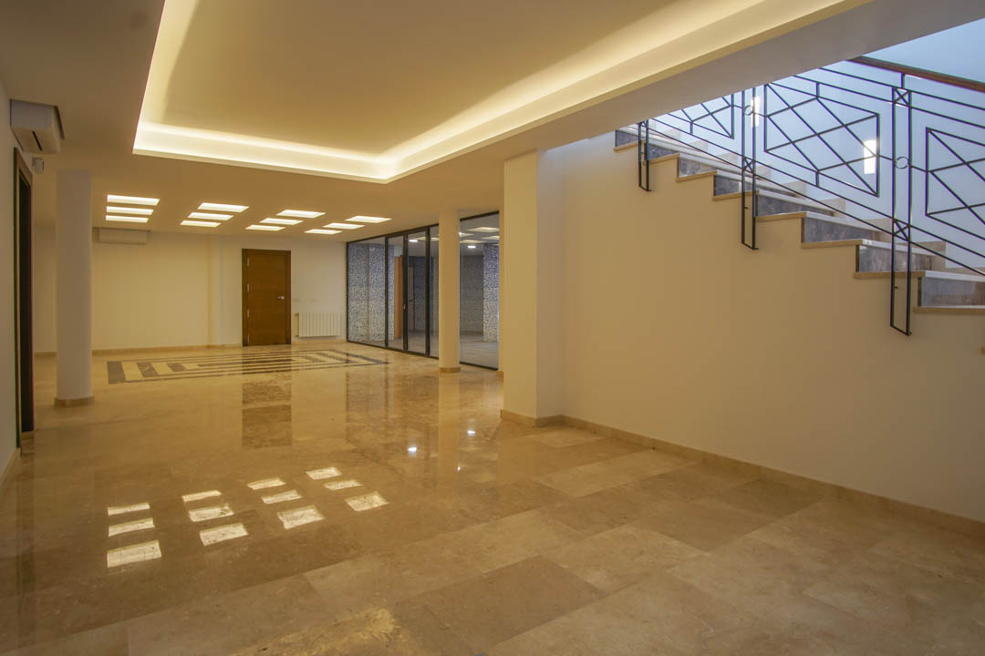 Ruime kelderlounge met gepolijste marmeren vloeren, geïntegreerde LED-plafondverlichting en glazen wanden die naar de wellnessruimte leiden.