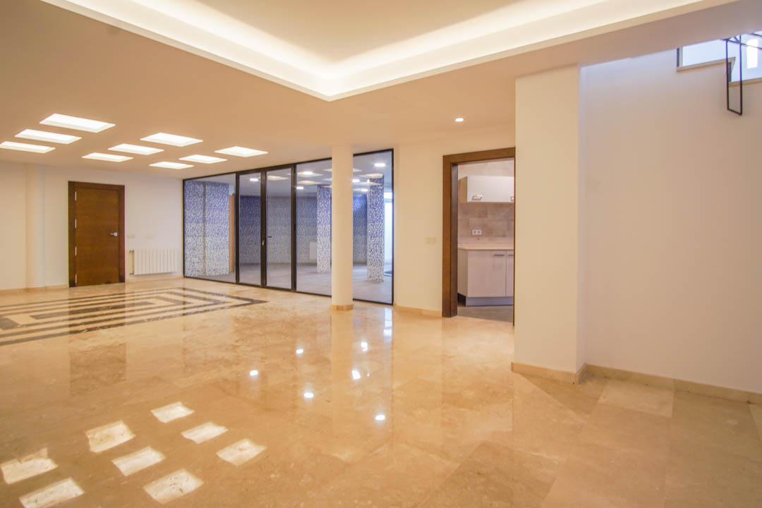 Ruime kelderverdieping met gepolijste marmeren vloeren, indirecte led-verlichting en een glazen wand die uitkijkt op een wellnessruimte met blauwe mozaïek.