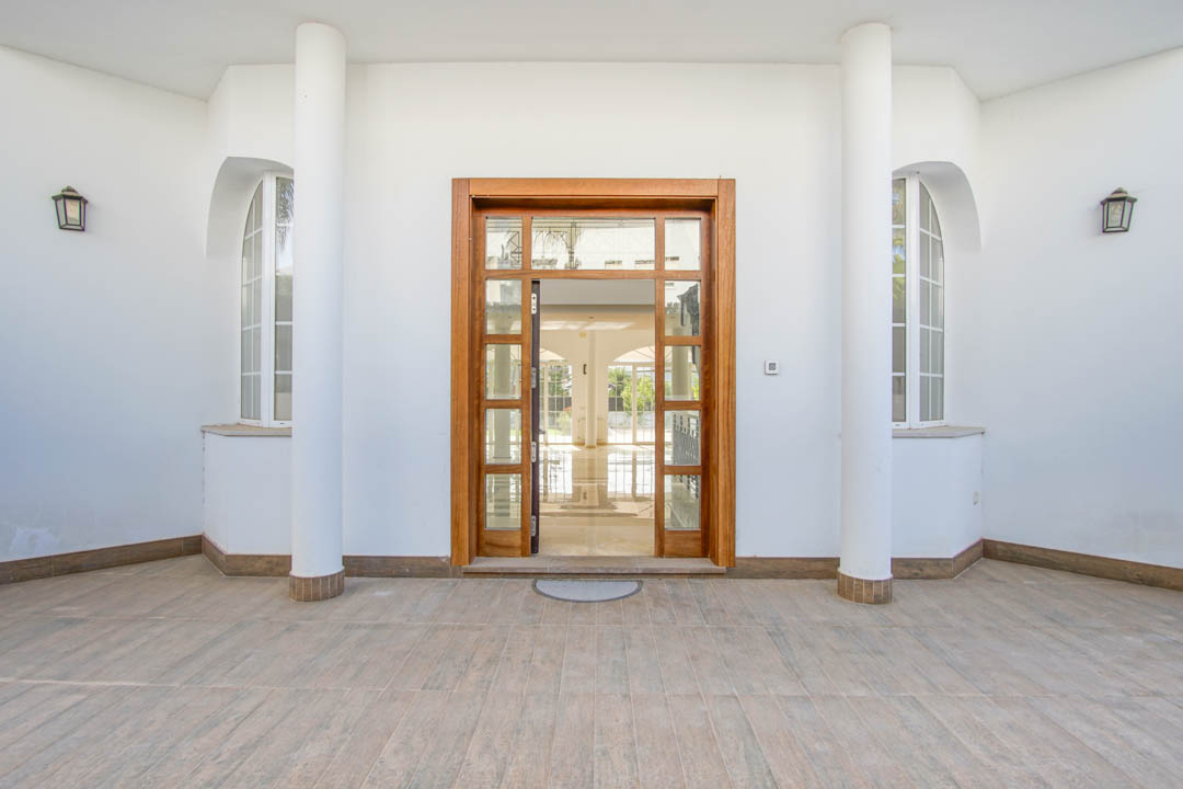 Symmetrische hoofdingang met een houten glazen deur, geflankeerd door witte zuilen en boogvensters met klassieke wandlantaarns.