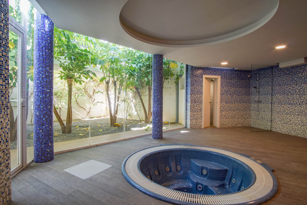 Wellnessruimte met ingebouwde ronde jacuzzi, blauwe mozaïekzuilen en glazen wanden die uitkijken op een besloten Engelse patio met volwassen bomen.