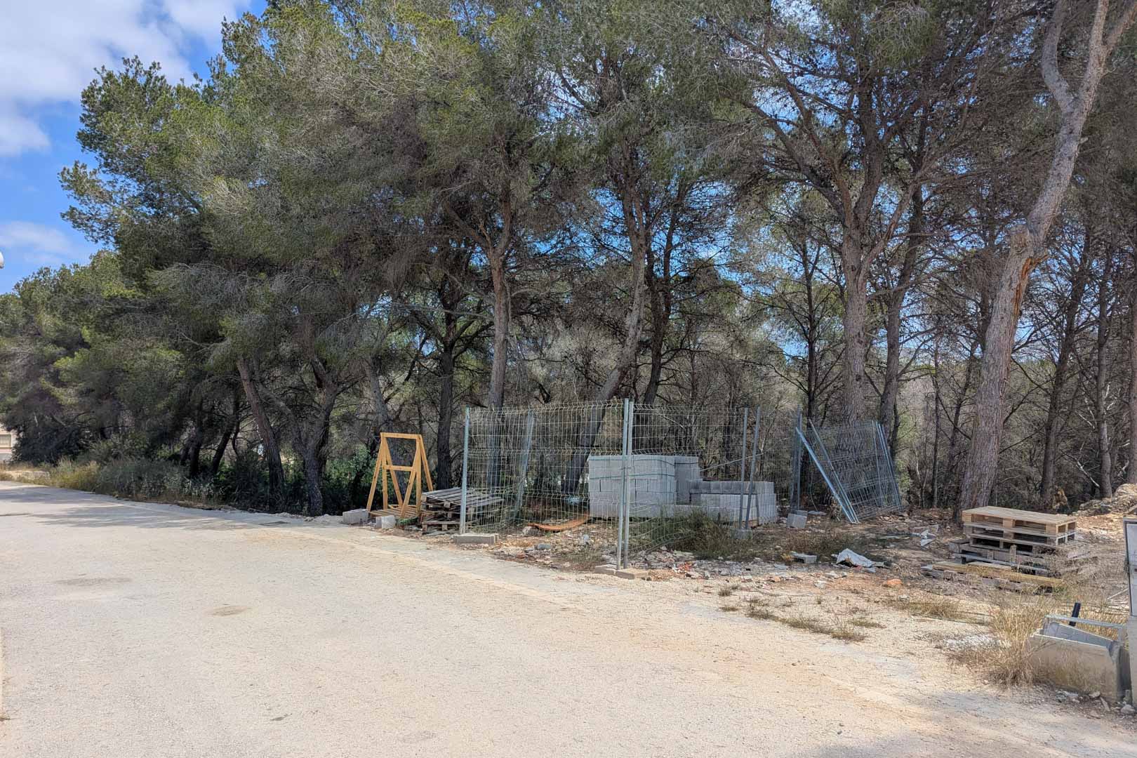 Bouwperceel met volwassen pijnbomen en eerste bouwmaterialen, gelegen aan een geasfalteerde weg in een rustige woonwijk.
