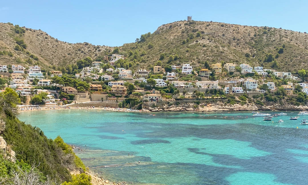 Panoramisch uitzicht op de baai van El Portet in Moraira, met turquoise wateren, voor anker liggende boten en witte villa's tegen de mediterrane heuvels.