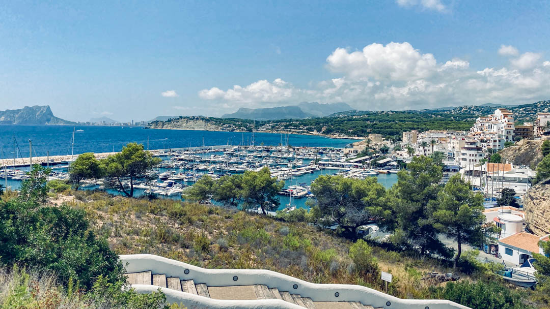 Panoramisch uitzicht op de jachthaven van Moraira en de Peñón de Ifach vanaf een witte trap in mediterrane stijl, omgeven door lokale kustvegetatie.