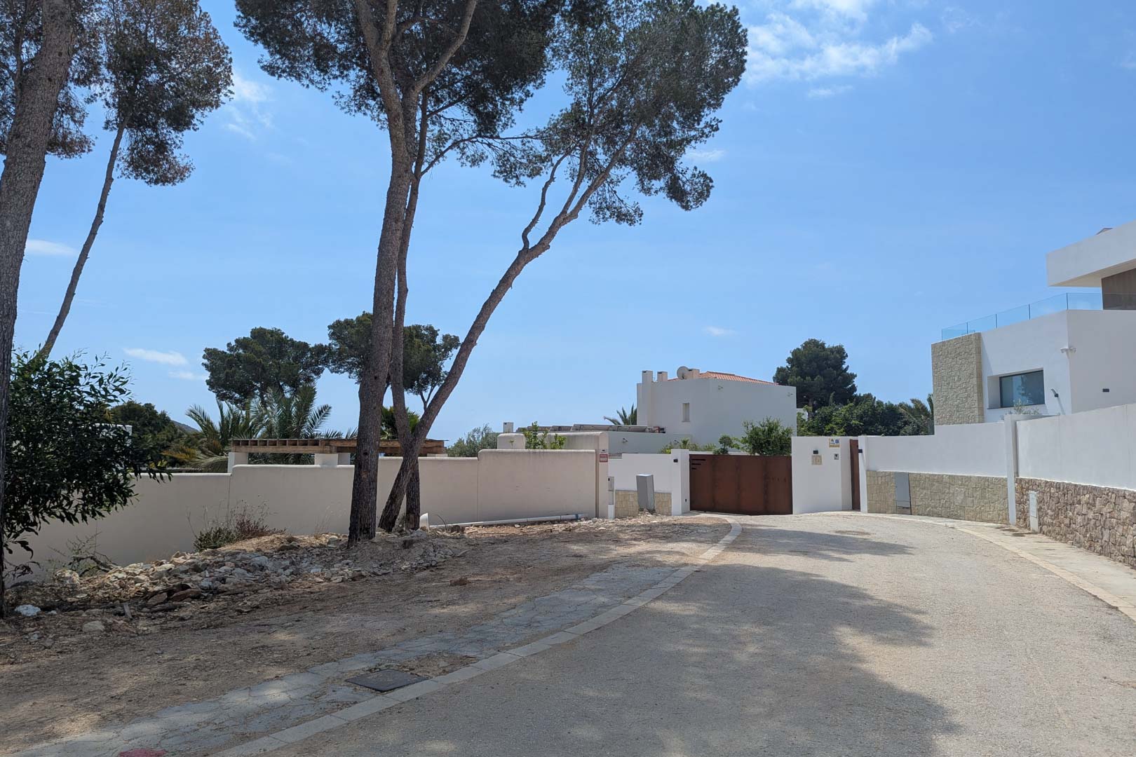 Toegang tot het perceel in een woonwijk in Moraira, met witte omheiningsmuren, volwassen pijnbomen en een verharde weg onder een helderblauwe lucht.