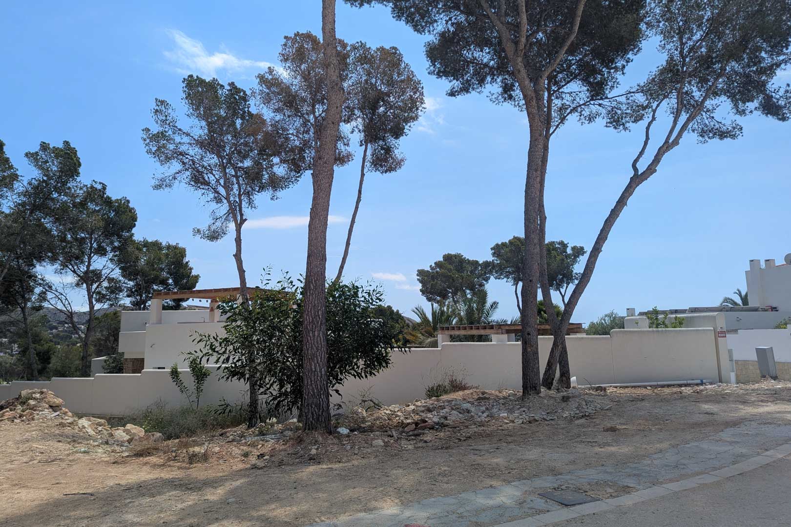 Zicht op het bouwperceel met volwassen pijnbomen en witte muren, klaar voor de bouw van de villa in Ibiza-stijl in Moraira.