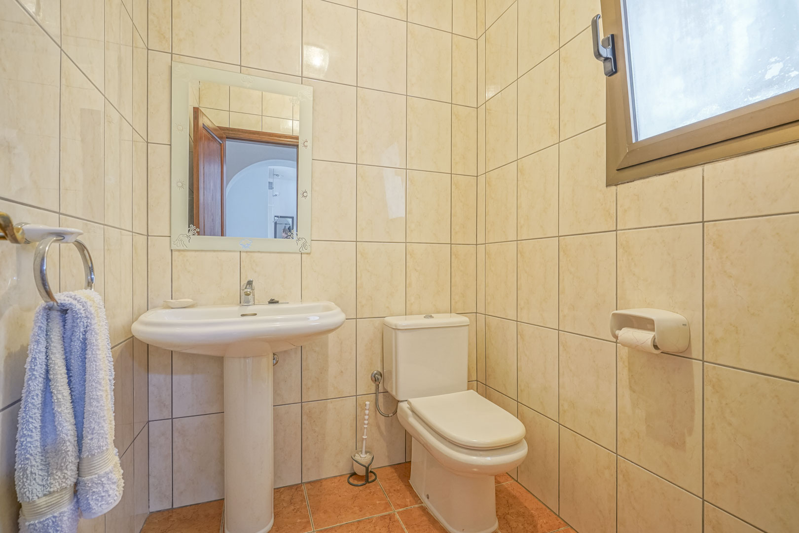 Badkamer met crèmekleurige tegels, een witte wastafel op voetstuk en een versierde spiegel.