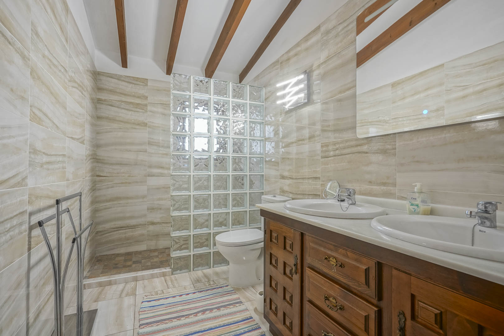 Badkamer met glazen bouwsteen douchewand, houten wastafelmeubel met dubbele wastafels en geaderde tegels.