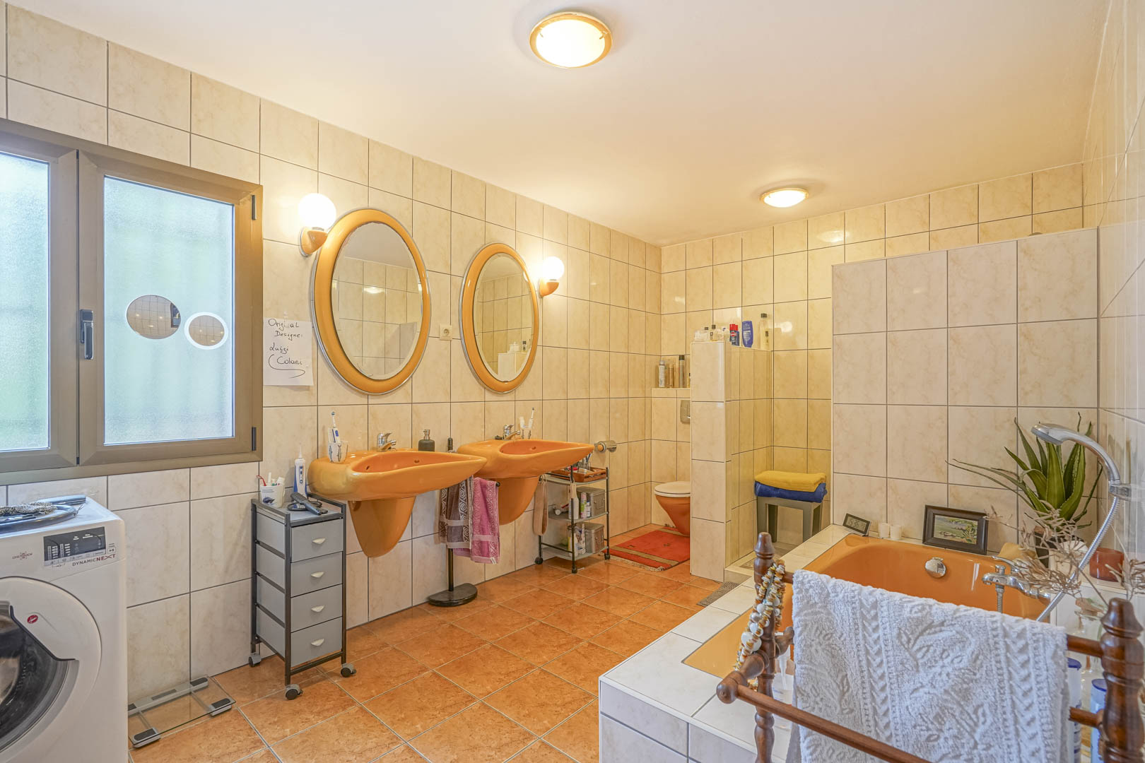 Badkamer met oranje wastafels en bijpassend bad, crèmekleurige tegels en een raam met natuurlijk licht.