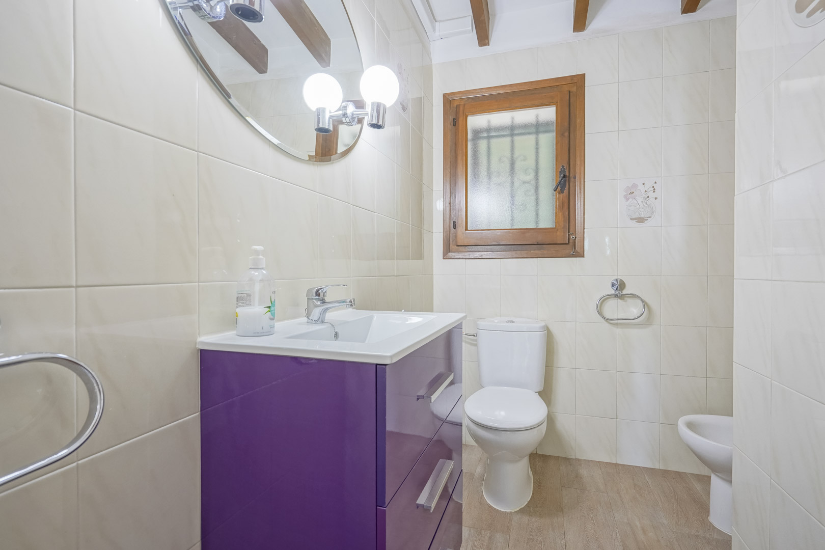 Close-up van een badkamer met crèmekleurige tegels, een paarse wastafel en een houten raam.