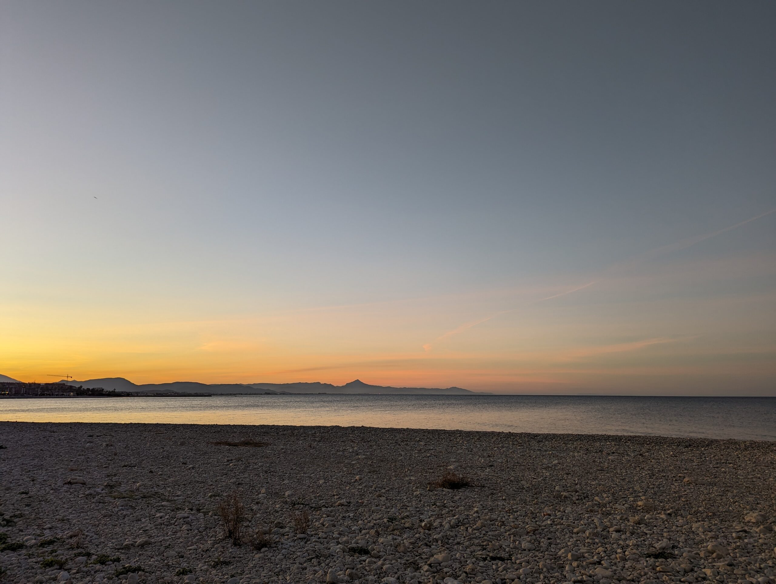 Uitzicht op de zonsopgang over een kiezelstrand met bergen op de achtergrond.
