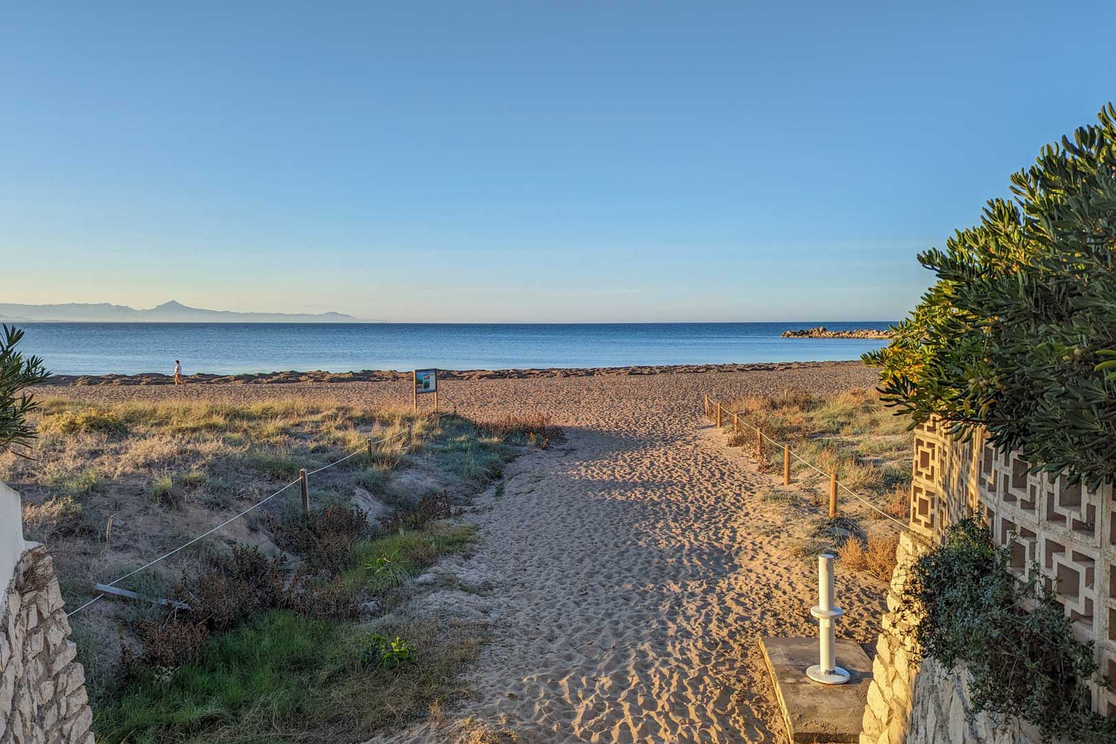 Uitzicht op zandstrand met afgezet pad, kustvegetatie en de blauwe zee onder een heldere hemel.