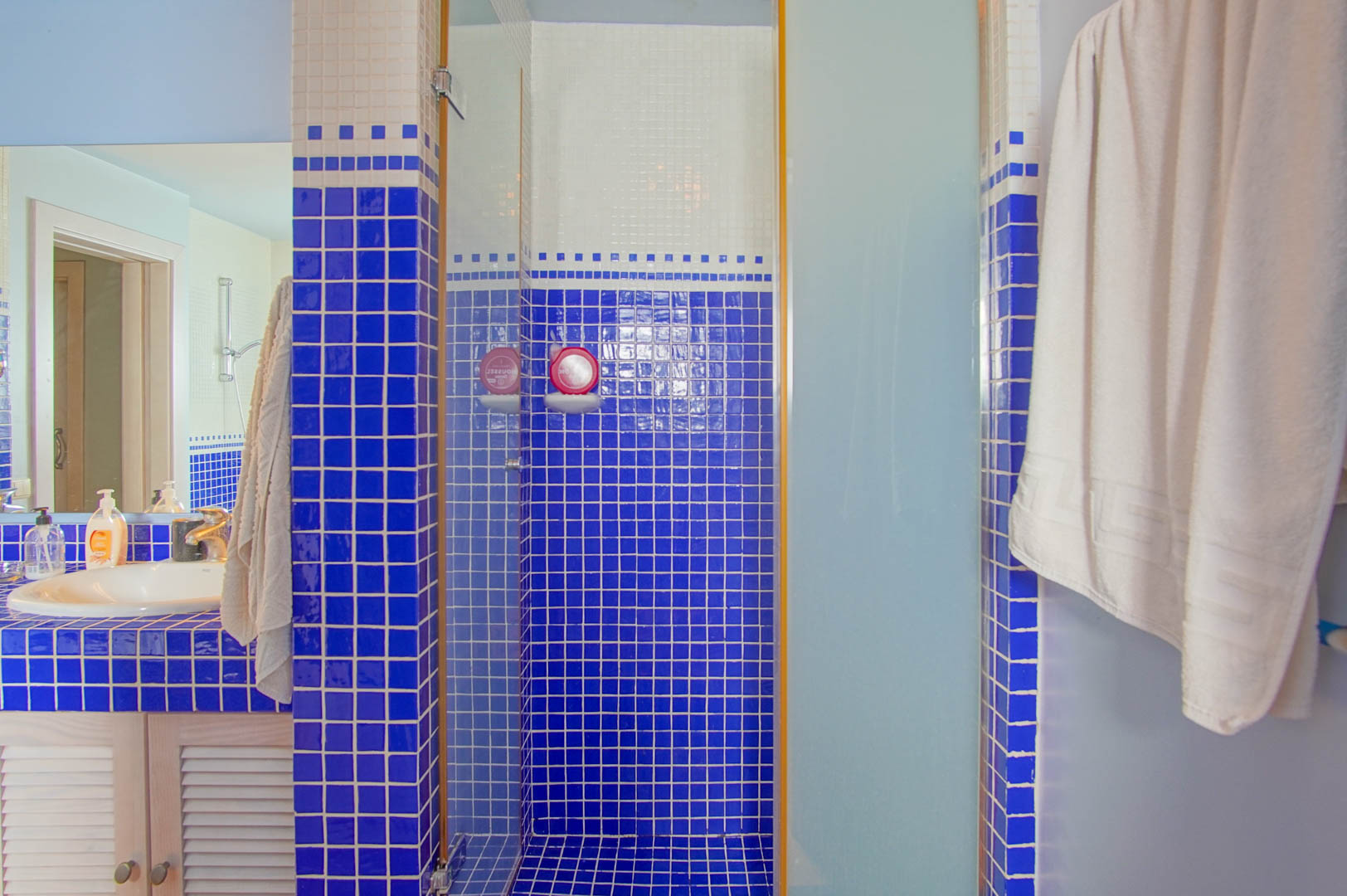 Badkamer met blauwe en witte mozaïektegels, een inloopdouche met glazen wand en een wastafelmeubel met houten lamellendeuren.