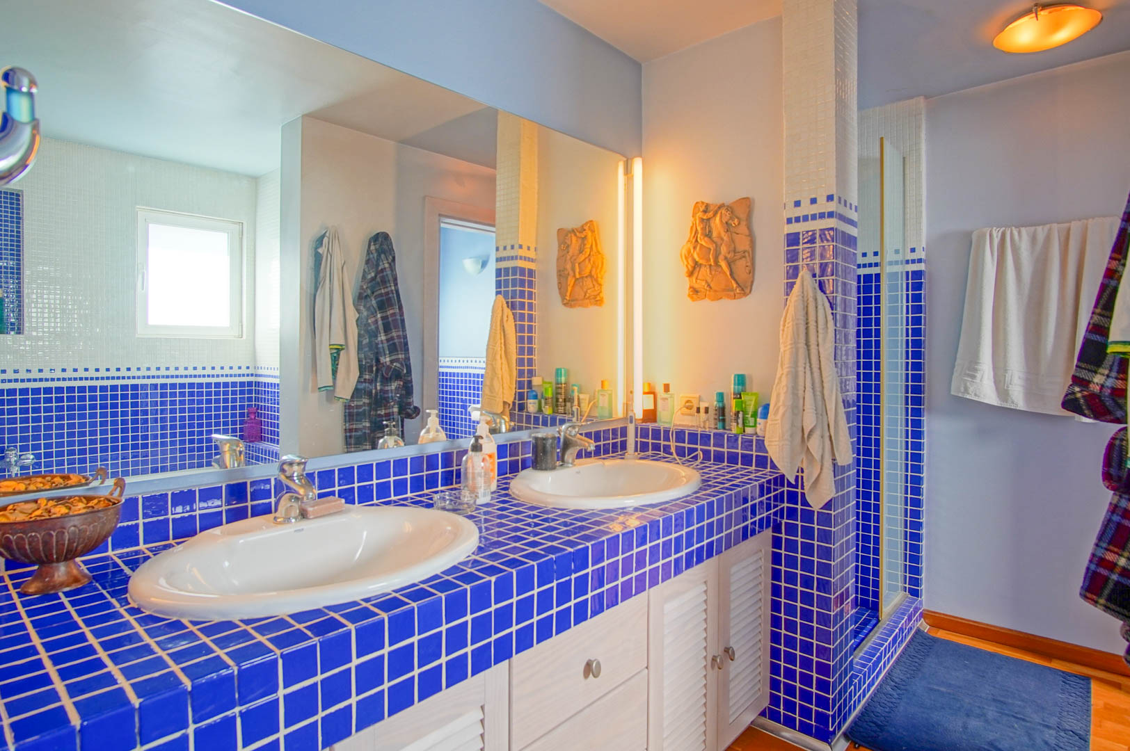 Badkamer met dubbele wastafel op een blauw betegeld werkblad en witte lamellenkastjes. Een grote spiegel reflecteert de doucheruimte en warme verlichting.