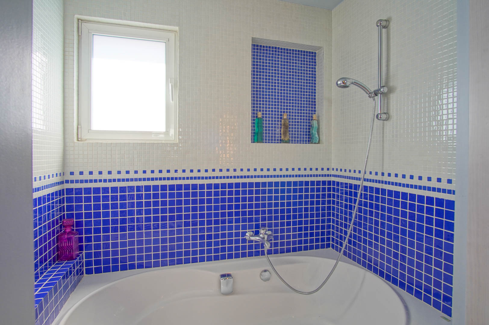 Badkamer met een ovaal bad en wanden van wit en kobaltblauw mozaïek. Voorzien van een ingebouwde nis voor toiletartikelen en een raam voor natuurlijk licht.