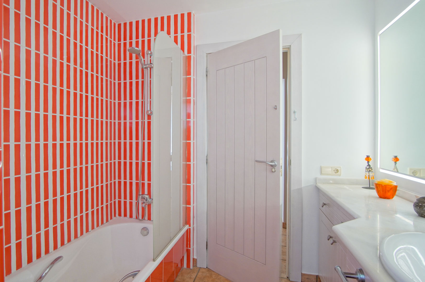 Badkamer met ligbad en glazen wand, opvallende oranje en witte verticale tegels, een wit meubel met marmeren werkblad en een spiegel met geïntegreerde verlichting.
