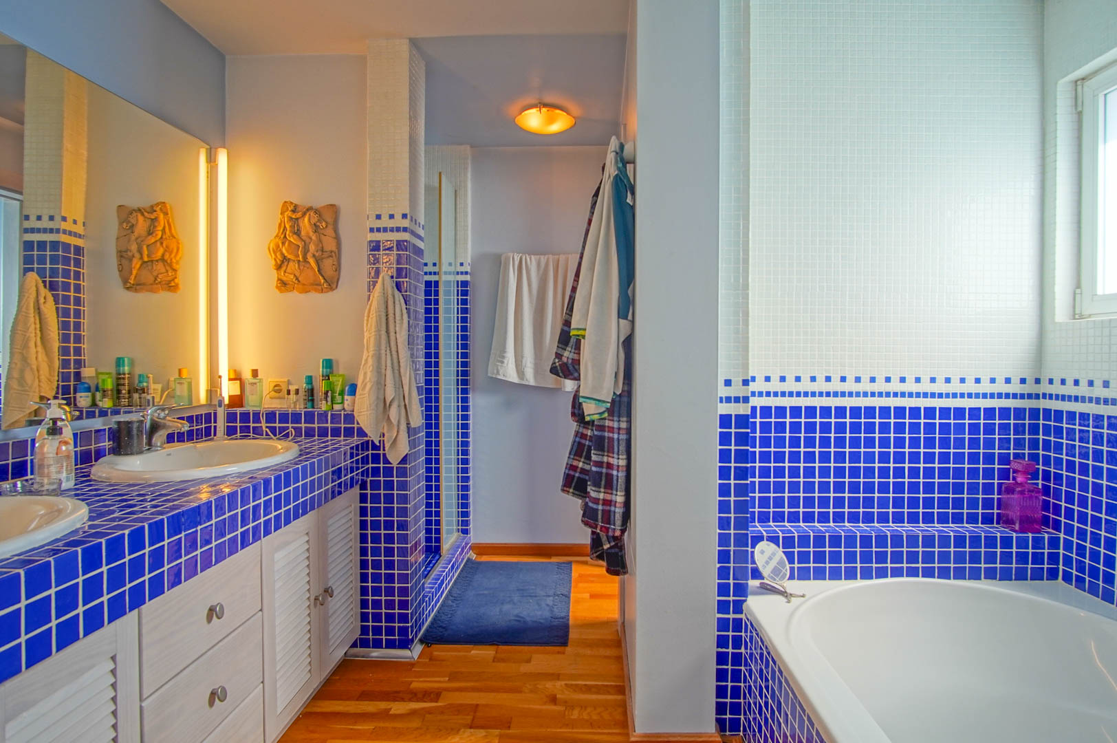 Badkamer voorzien van blauw-witte mozaïektegels, dubbele wastafel met lamellenkastjes, houten vloer en een ingebouwd ligbad naast een raam voor natuurlijk licht.