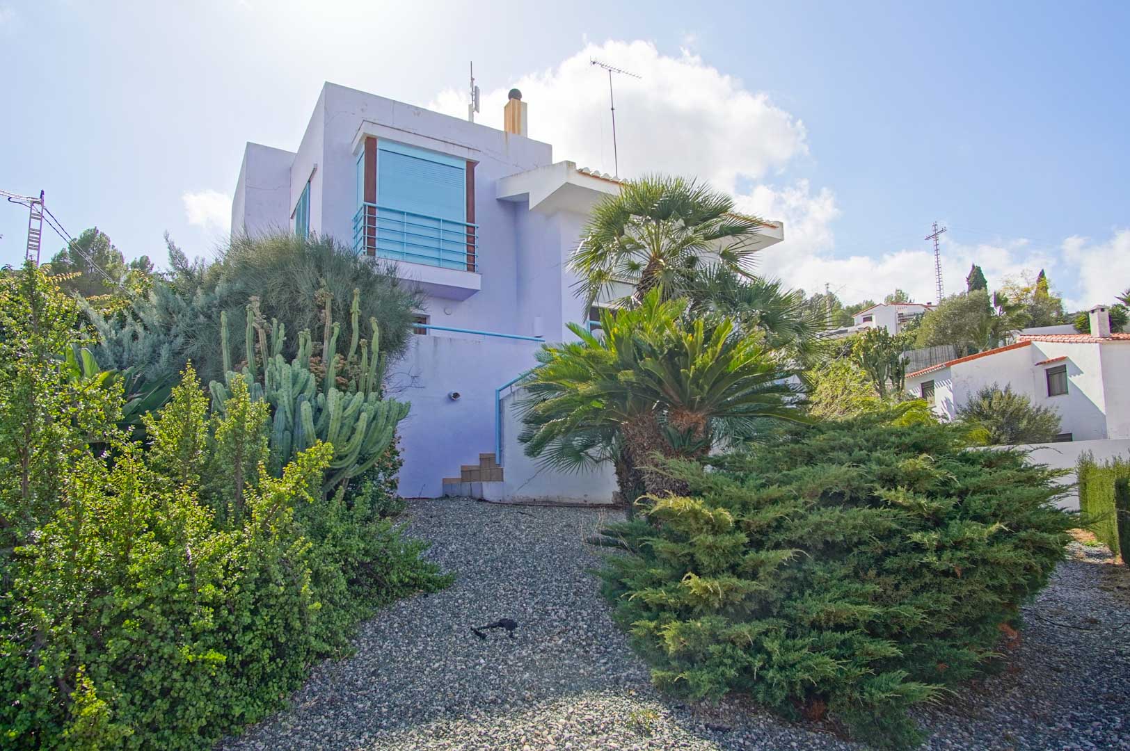 Buitenaanzicht van de lavendelkleurige gevel, geïntegreerd in een mediterrane tuin met cactussen, palmbomen en struiken op een grindpad.