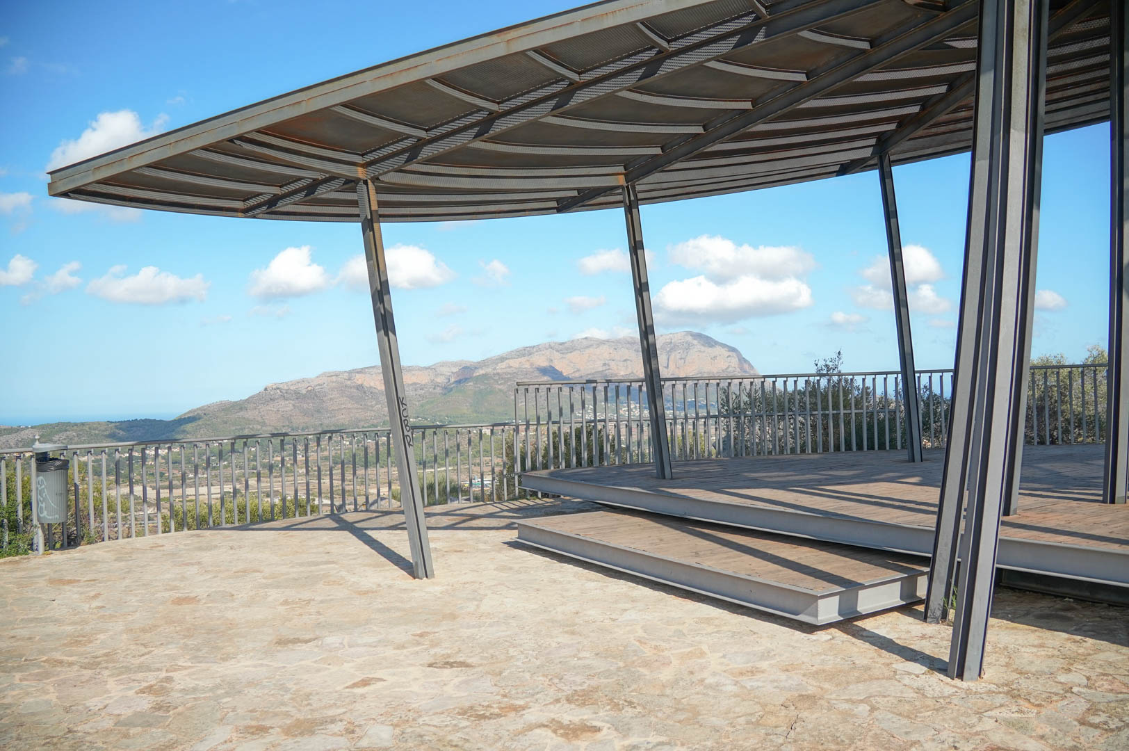 Buitenuitkijkpunt met een moderne staalconstructie en houten terras, met panoramisch uitzicht op de bergen en de vallei.