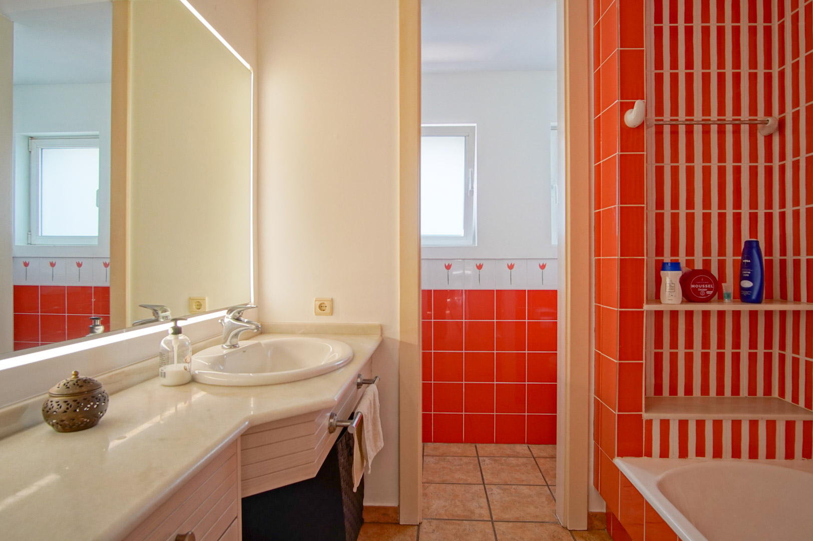 Functionele badkamer met marmeren werkblad, verlichte spiegel en levendige oranje tegels. Voorzien van een badgedeelte met ingebouwde schappen en natuurlijk licht.