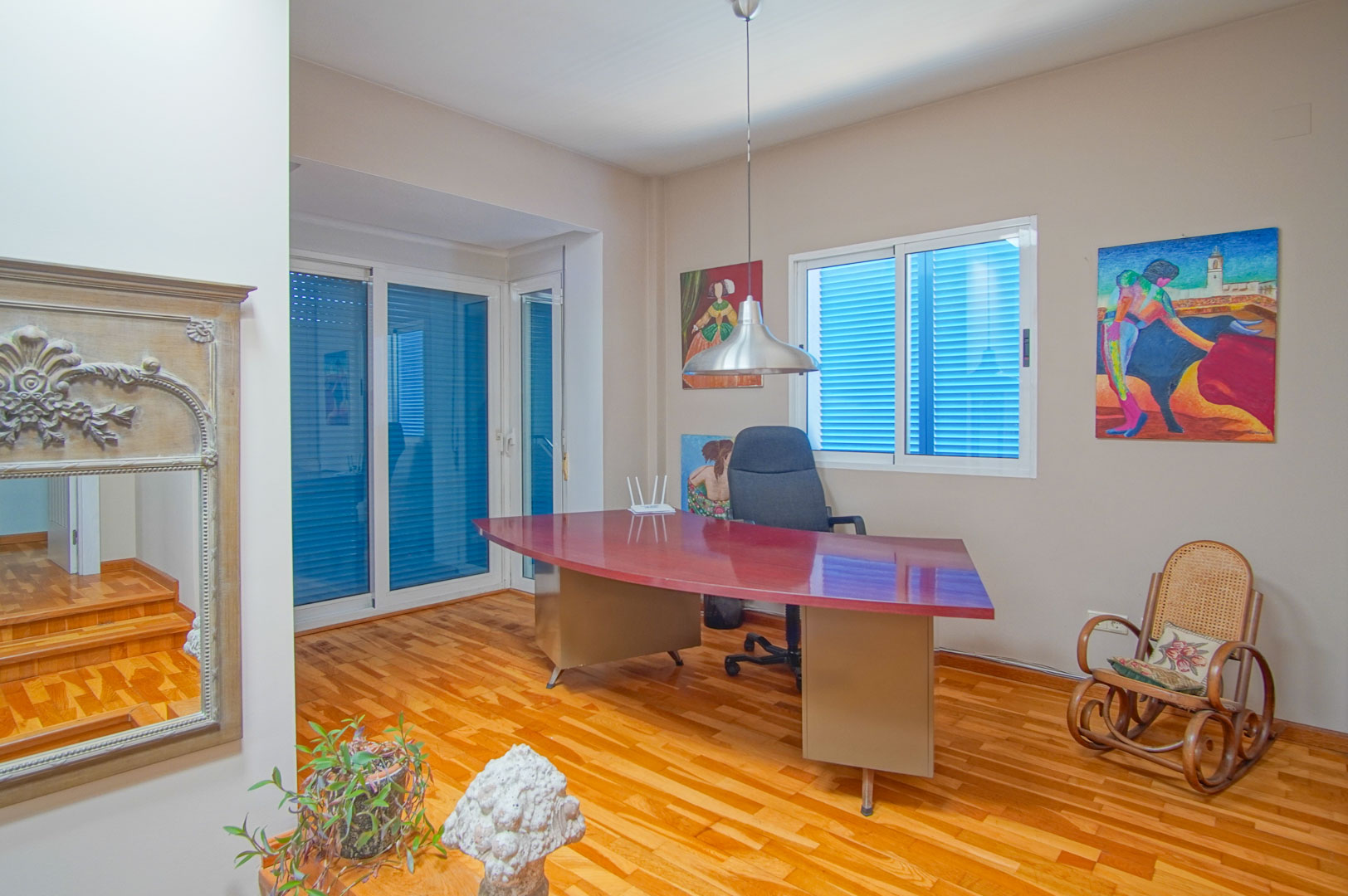 Licht kantoor met parketvloer, een groot gebogen bordeauxrood bureau en ramen met traditionele blauwe luiken voor natuurlijk licht.