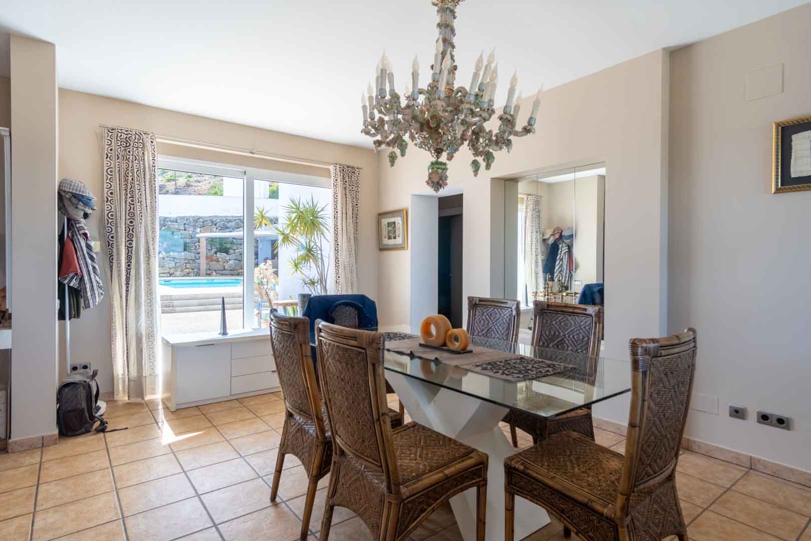 Lichte eetkamer met glazen tafel, rotan stoelen en een klassieke kroonluchter, met direct uitzicht op het zwembad via de grote raampartij.