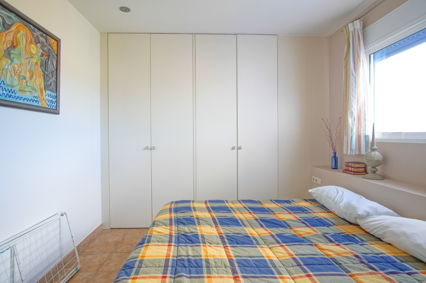 Lichte slaapkamer voorzien van grote witte inbouwkasten, een rustieke tegelvloer en een zijraam voor een optimale natuurlijke lichtinval.