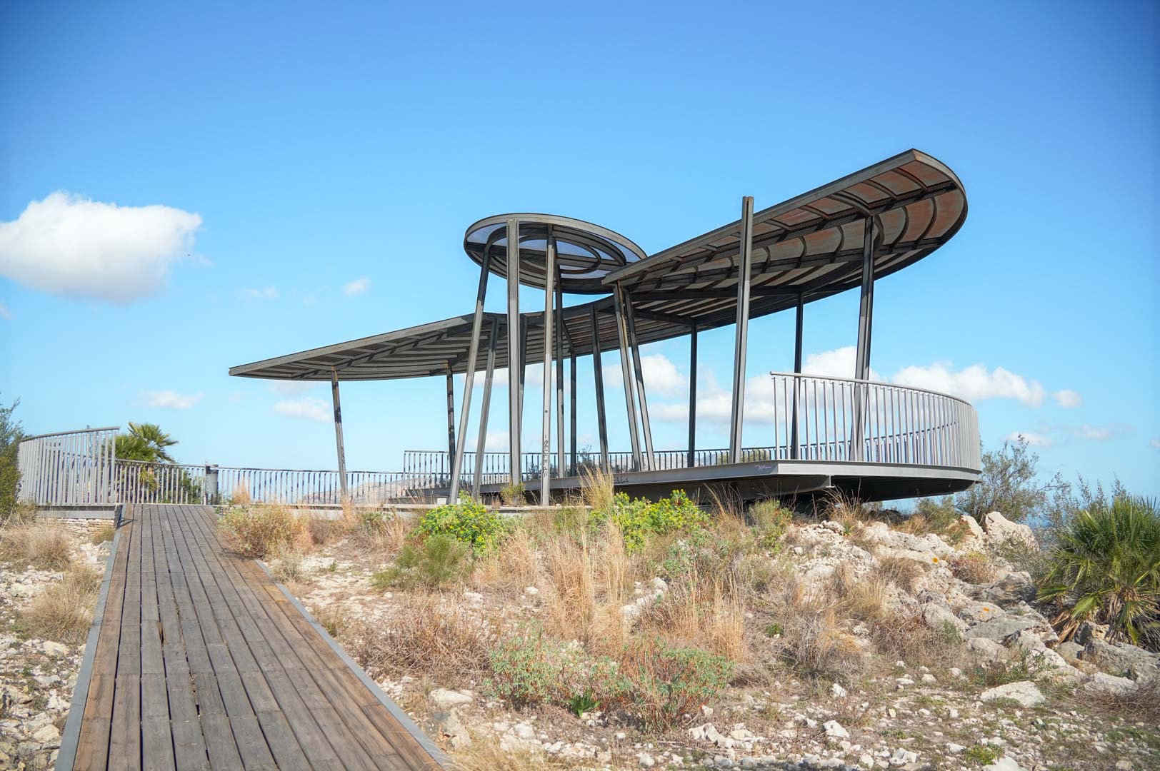Modern uitkijkpunt met een metalen structuur en gebogen dak, bereikbaar via een houten loopbrug in een natuurlijk mediterraan landschap.