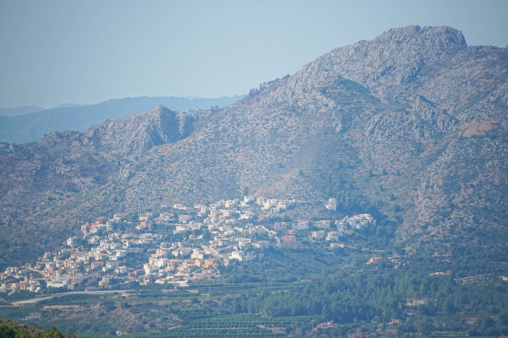 Panoramisch uitzicht op de bergketen en de vallei, met een woonwijk op de helling boven uitgestrekte groene mediterrane landbouwvelden.