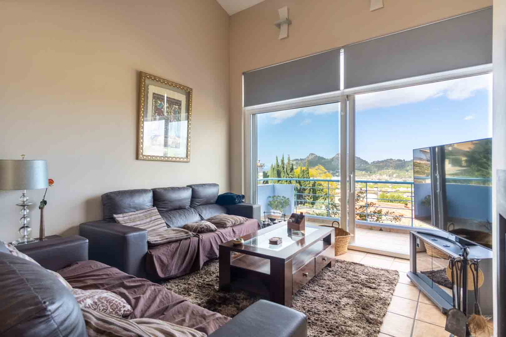 Woonkamer met hoge plafonds, donkere banken en een houten salontafel. Grote glazen schuifdeuren bieden toegang tot het balkon met panoramisch uitzicht op de bergen.